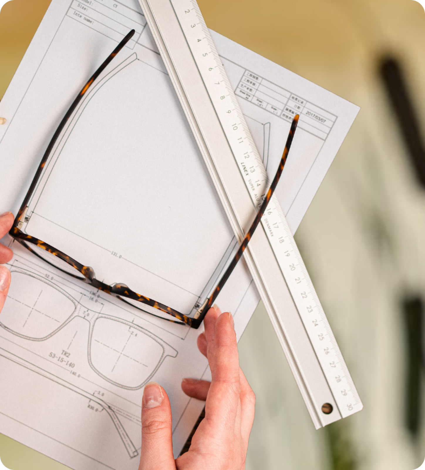 Hands measuring glasses with a paper ruler on top of a design sketch and tortoiseshell frames.