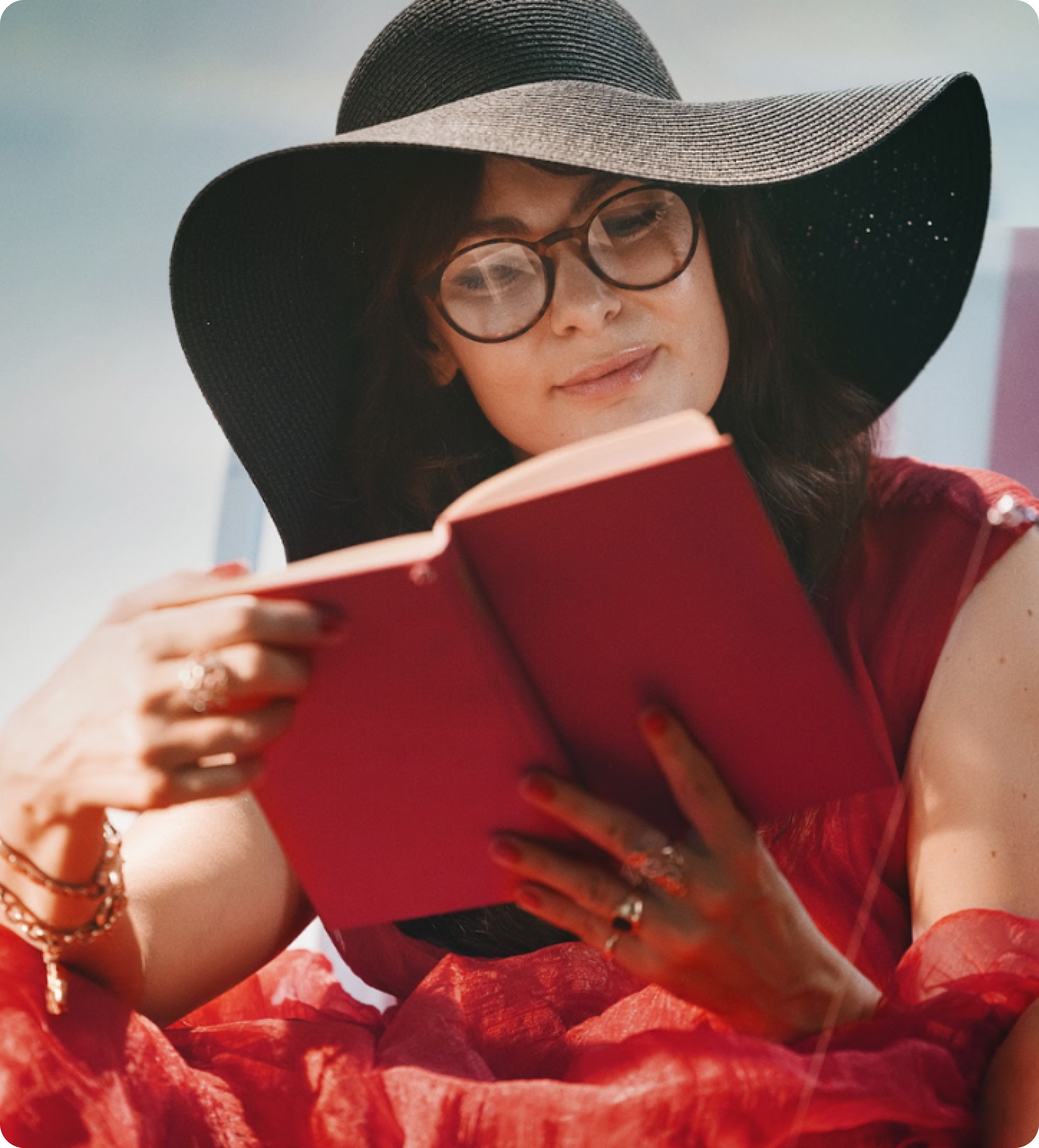 Woman in red dress with black hat and glasses reading a red book outdoors, smiling contently.