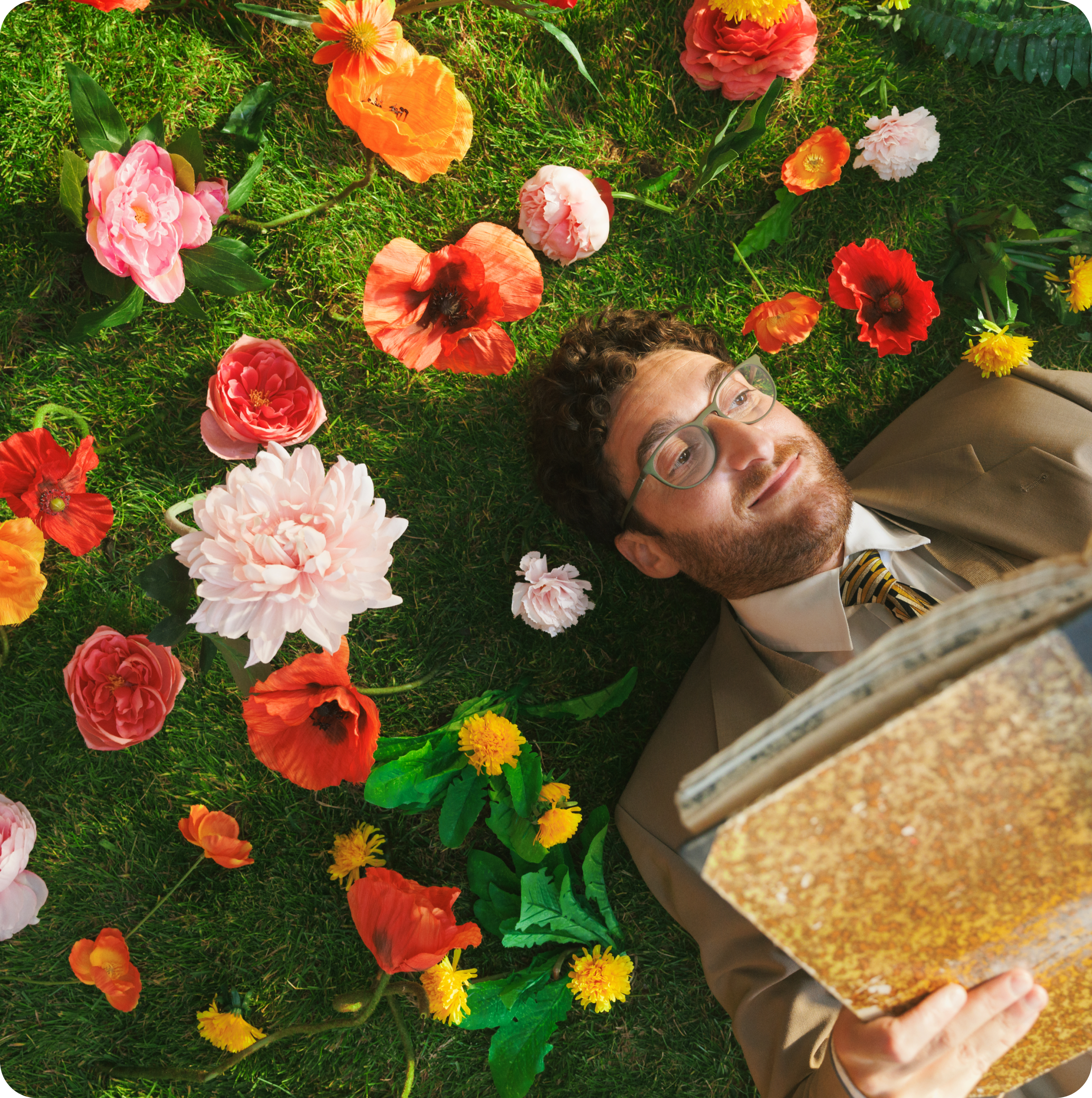 Man in glasses lying on grass with colorful flowers, reading a book.
